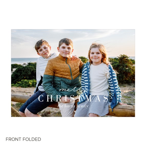 Christmas photo card: three children on a wooden rail at the beach with ocean and dunes; white 'Merry Christmas' text overlay.