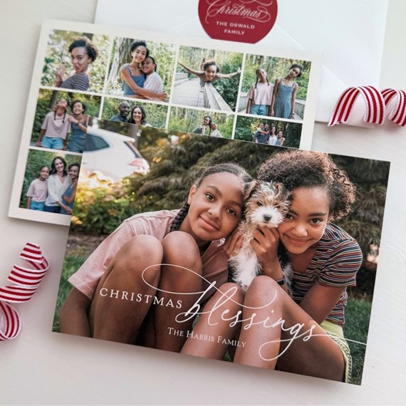Holiday photo card with two girls and a small puppy on the front, collage card and envelope visible behind, white script greeting across the image.