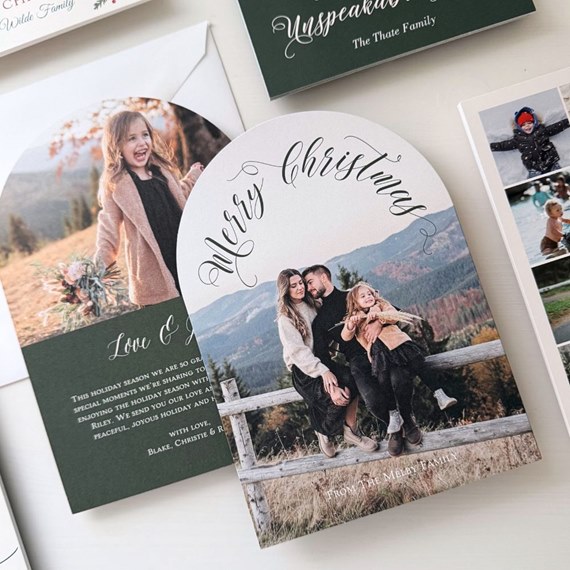Arch-shaped Christmas photo card with cursive 'Merry Christmas' and family portrait on a wooden fence in a mountain setting