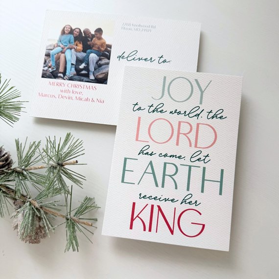 Christmas postcard with large multicolor typographic message ('Joy...King') on textured white card and a family photo on the reverse, shown with pine sprigs.