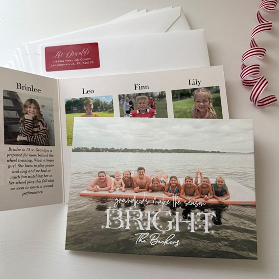 Folded holiday photo card with children on a paddleboard in a lake, white script title on front and an inside collage of individual portraits; envelopes and a red address label shown.