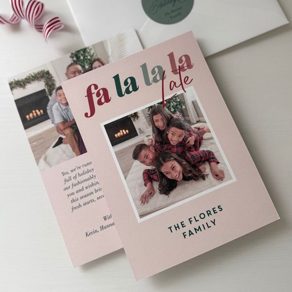 Pink folded Christmas photo card with "fa la la late" headline and white-bordered family photo, shown with envelope.